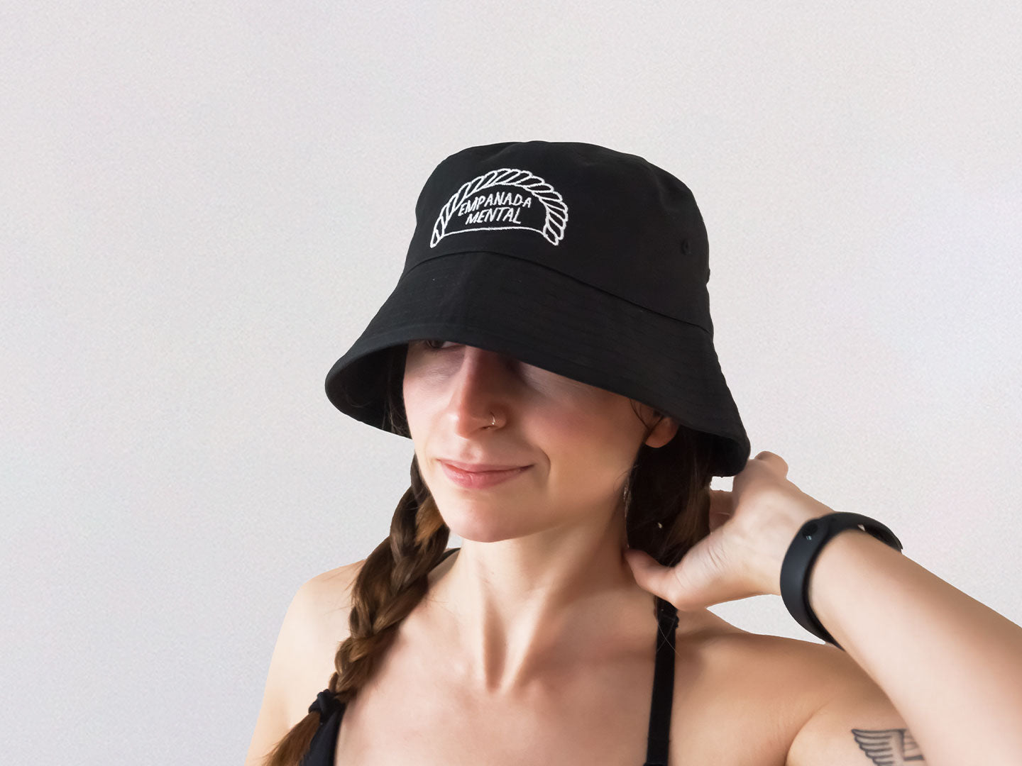 Chica con trenzas que lleva un gorro de pescador bucket hat negro con un bordado de una ilustración de una empanadilla en la que pone "empanada mental" de la marca Lola Pistola