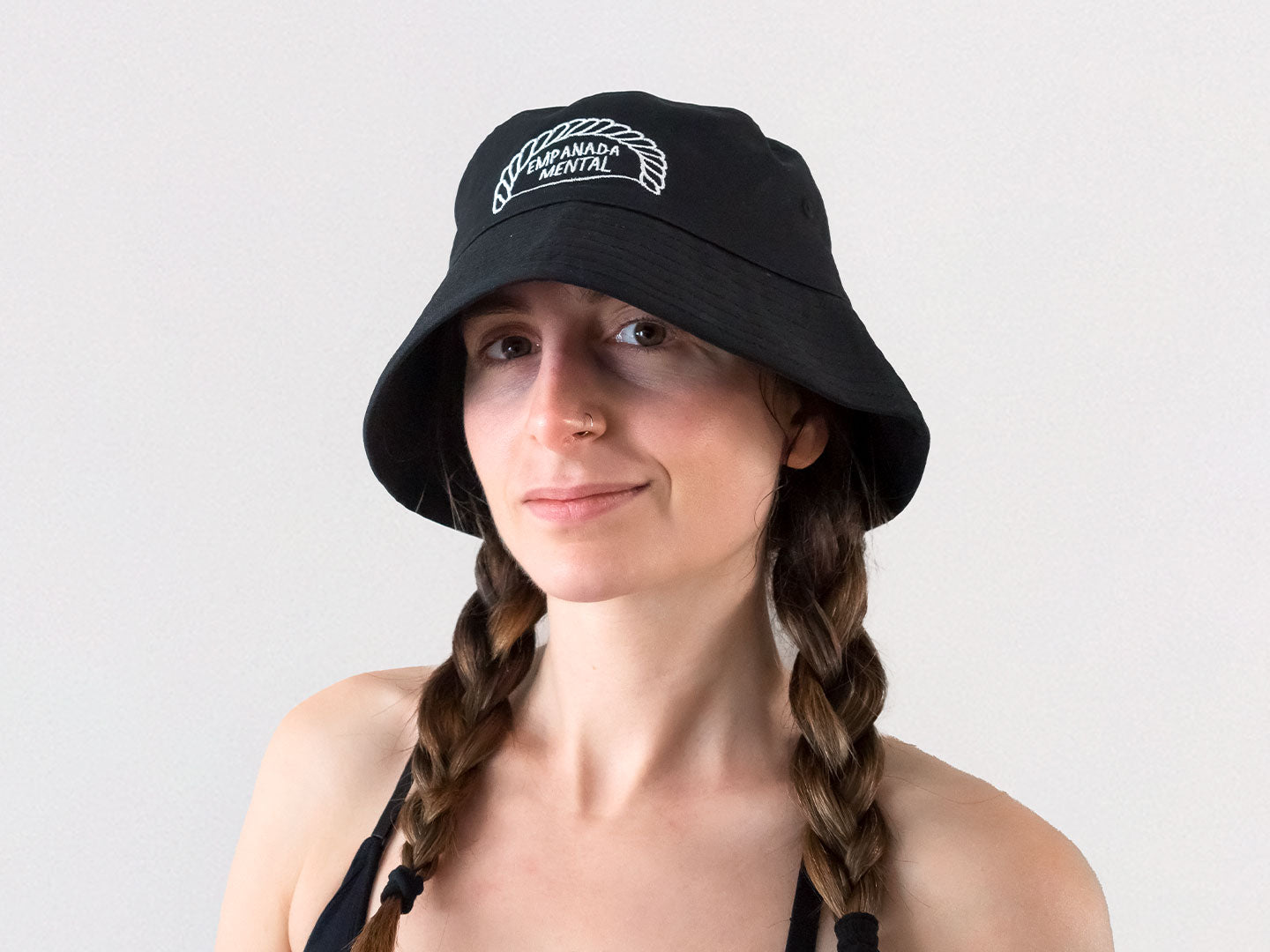 Chica con trenzas que lleva un gorro de pescador bucket hat negro con un bordado de una ilustración de una empanadilla en la que pone "empanada mental"